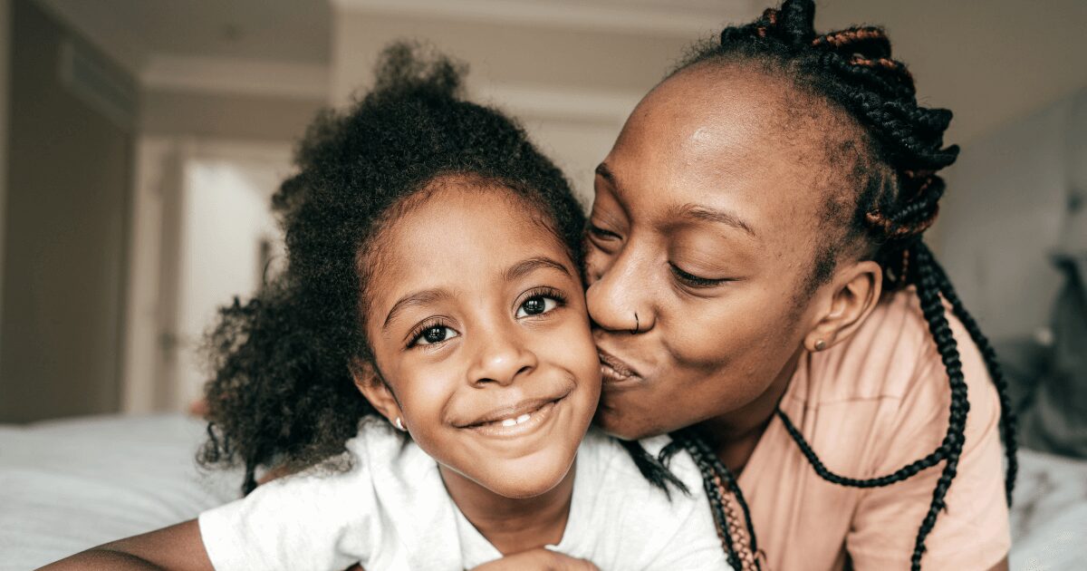 A woman smiles as she kisses her child who looks at the camera